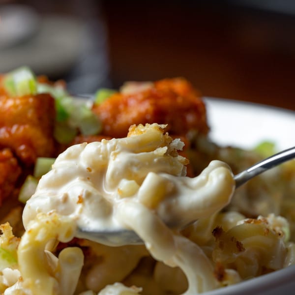 A spoon lifts creamy mac and cheese with crispy breadcrumbs, alongside buffalo chicken and celery.