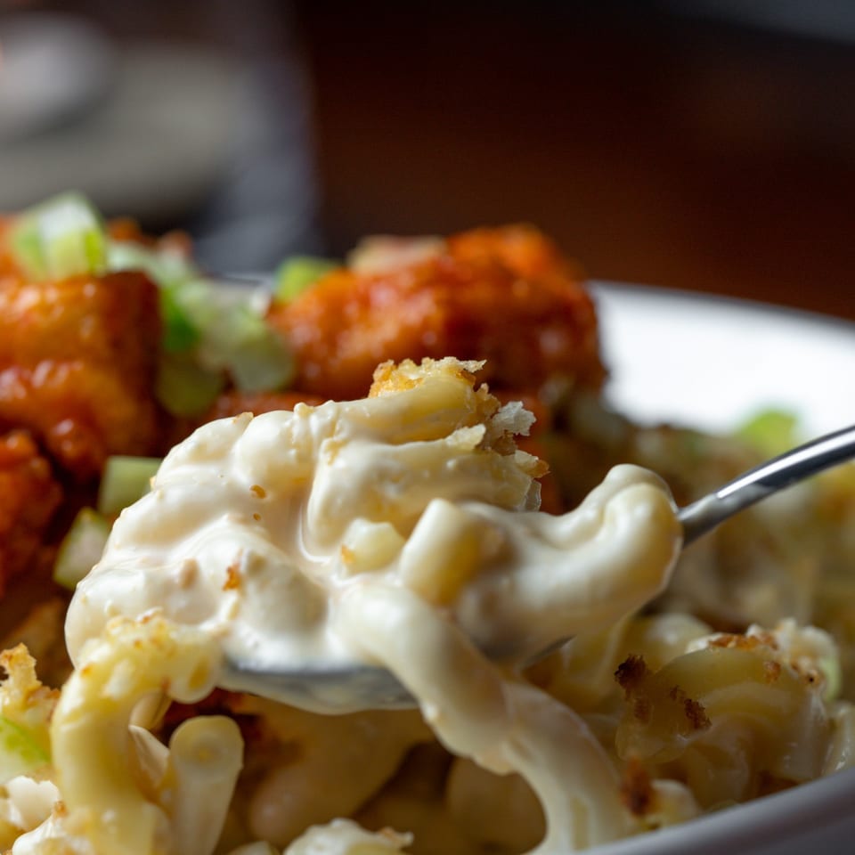 A spoon lifts creamy mac and cheese with crispy breadcrumbs, alongside buffalo chicken and celery.