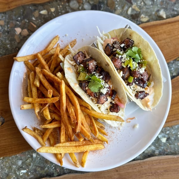 Two loaded tacos with seasoned fries on a white plate, set on a wooden and stone table.