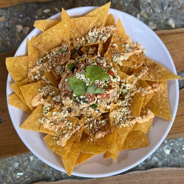 Nachos with creamy sauce, crumbled cheese, and cilantro in a white bowl on a wooden table.