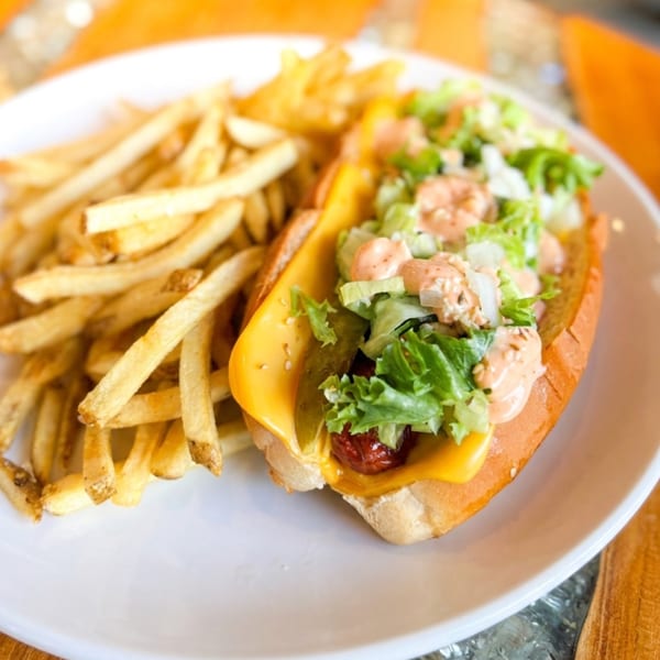 Hot dog with cheese, pickles, lettuce, and sauce, served with a side of golden french fries.
