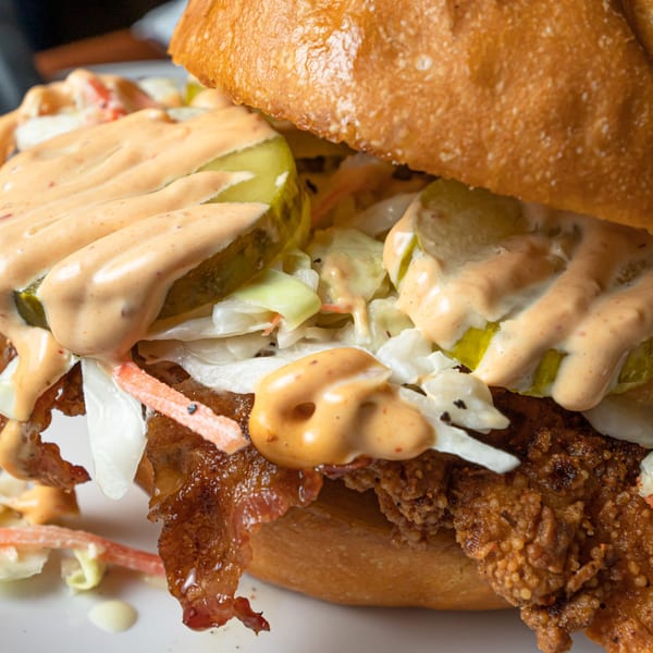 Close-up of a fried chicken sandwich with pickles, coleslaw, bacon, and special sauce on a bun.