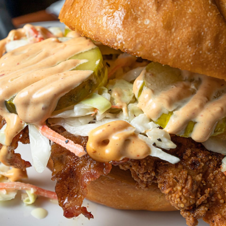 Close-up of a fried chicken sandwich with pickles, coleslaw, bacon, and special sauce on a bun.